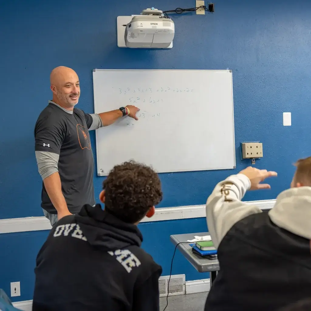A man points to a whiteboard in front of a class.