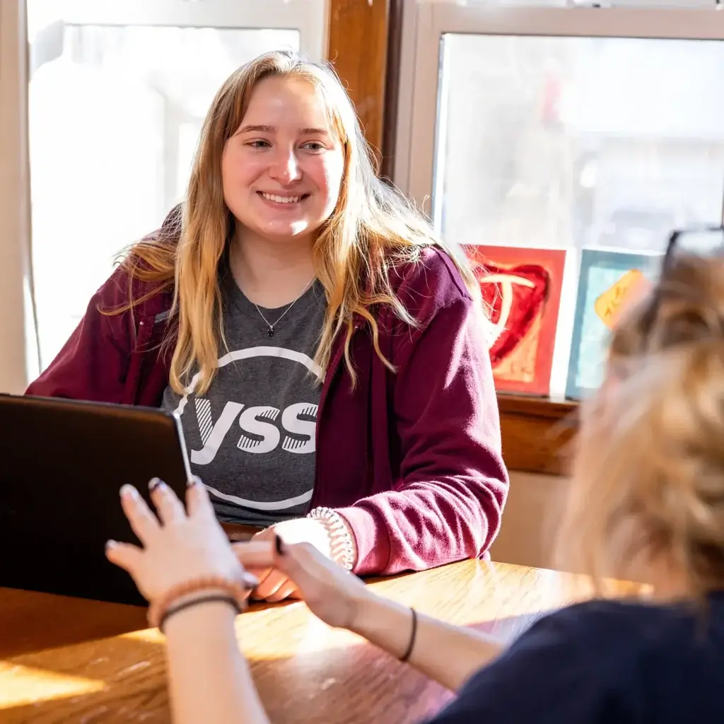 A woman wearing a gray YSS shirt sits at a table with a laptop, smiling at another woman who is partially out of frame.
