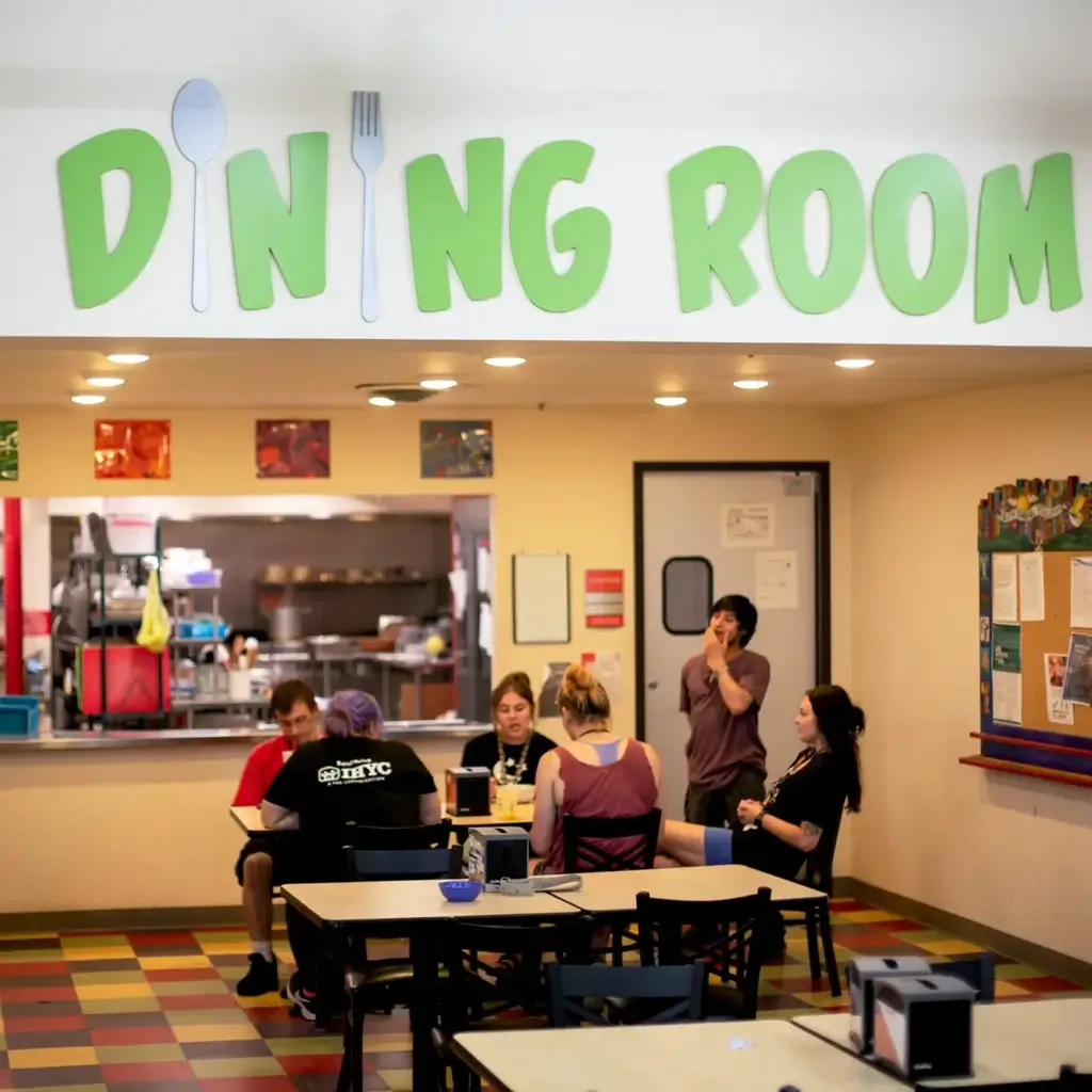 A group of teens eating at a cafeteria table. There is a green sign above that says "Dining Room"