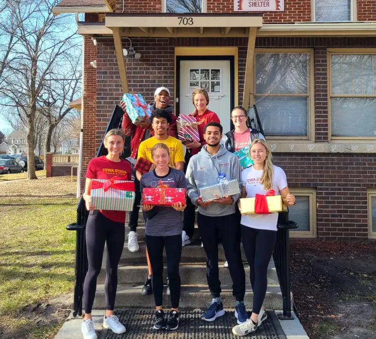 A group of Iowa State college students holding wrapped gifts on the front steps of a house.