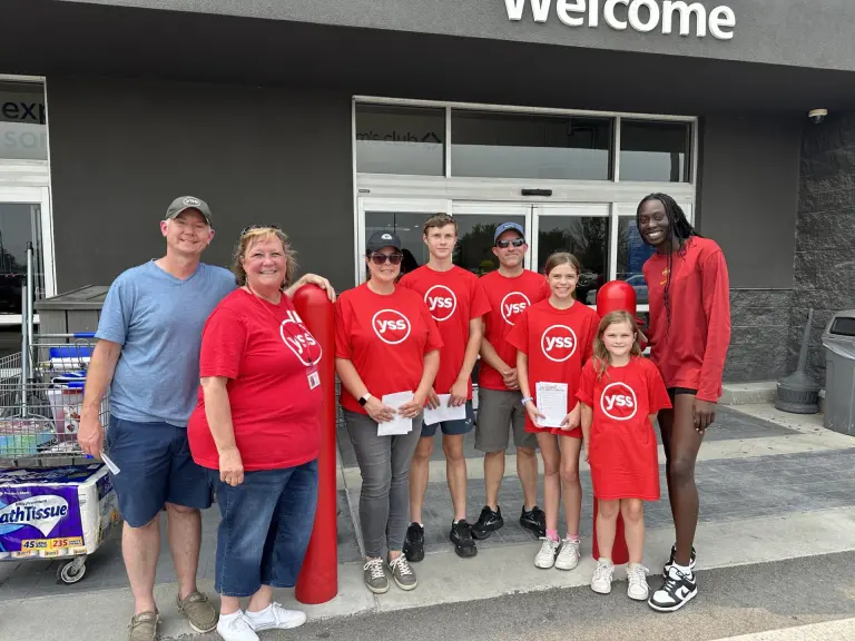 A group of adults and children, most wearing red YSS shirts, standing in front of a building