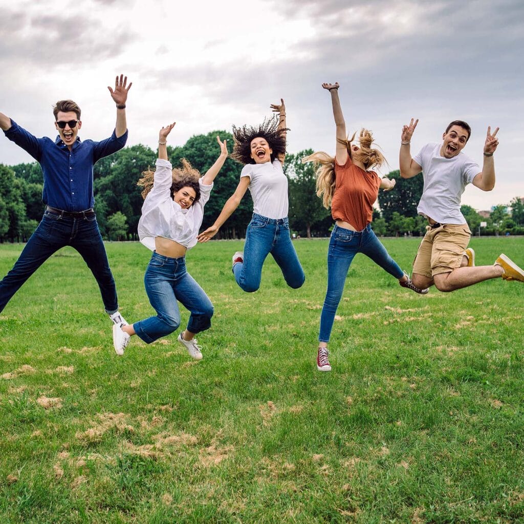 Happy group of teens jumping with their arms up outside in a grass field.