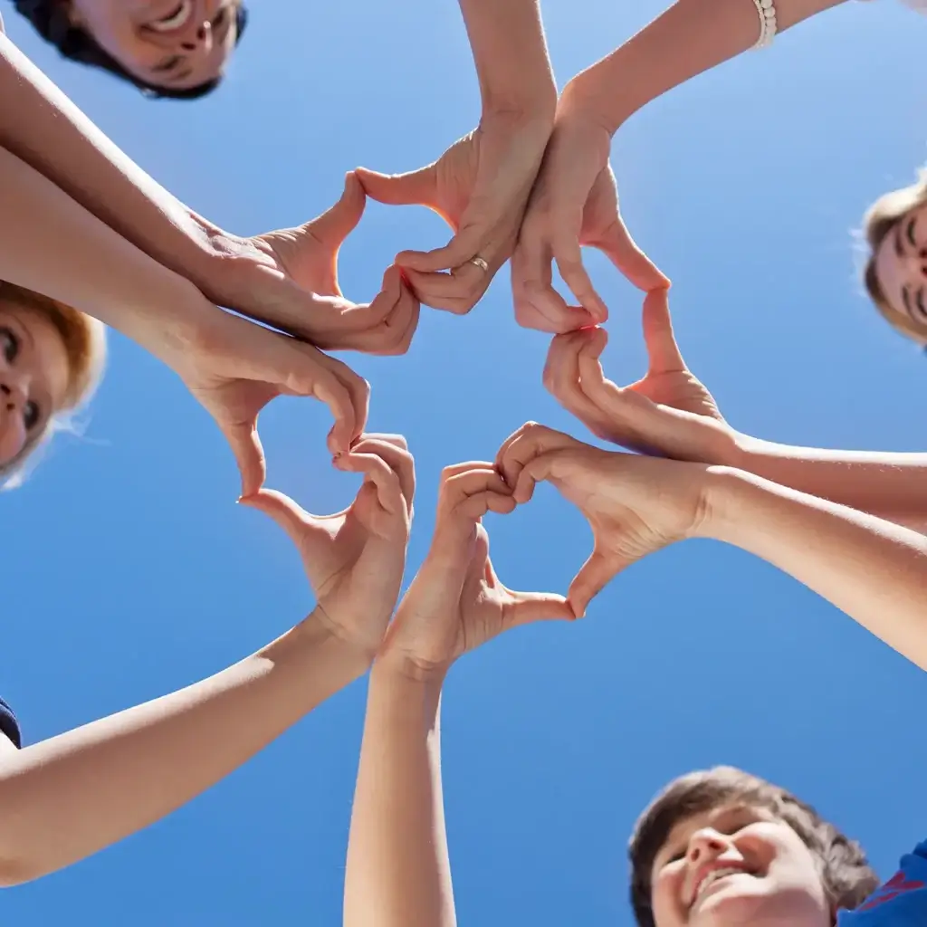 Kids standing in a circle making hearts with their hands. There is a clear blue sky in the background.