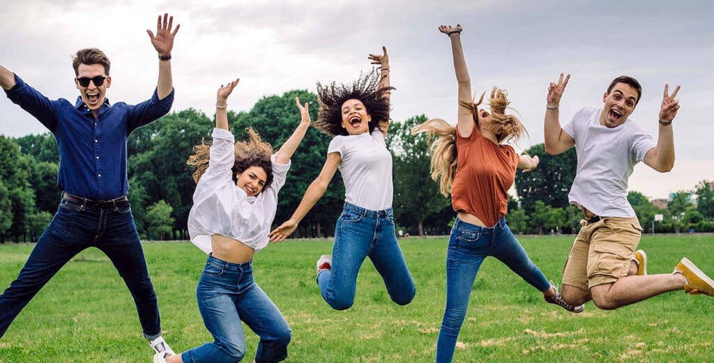 Happy group of teens jumping with their arms up outside in a grass field.