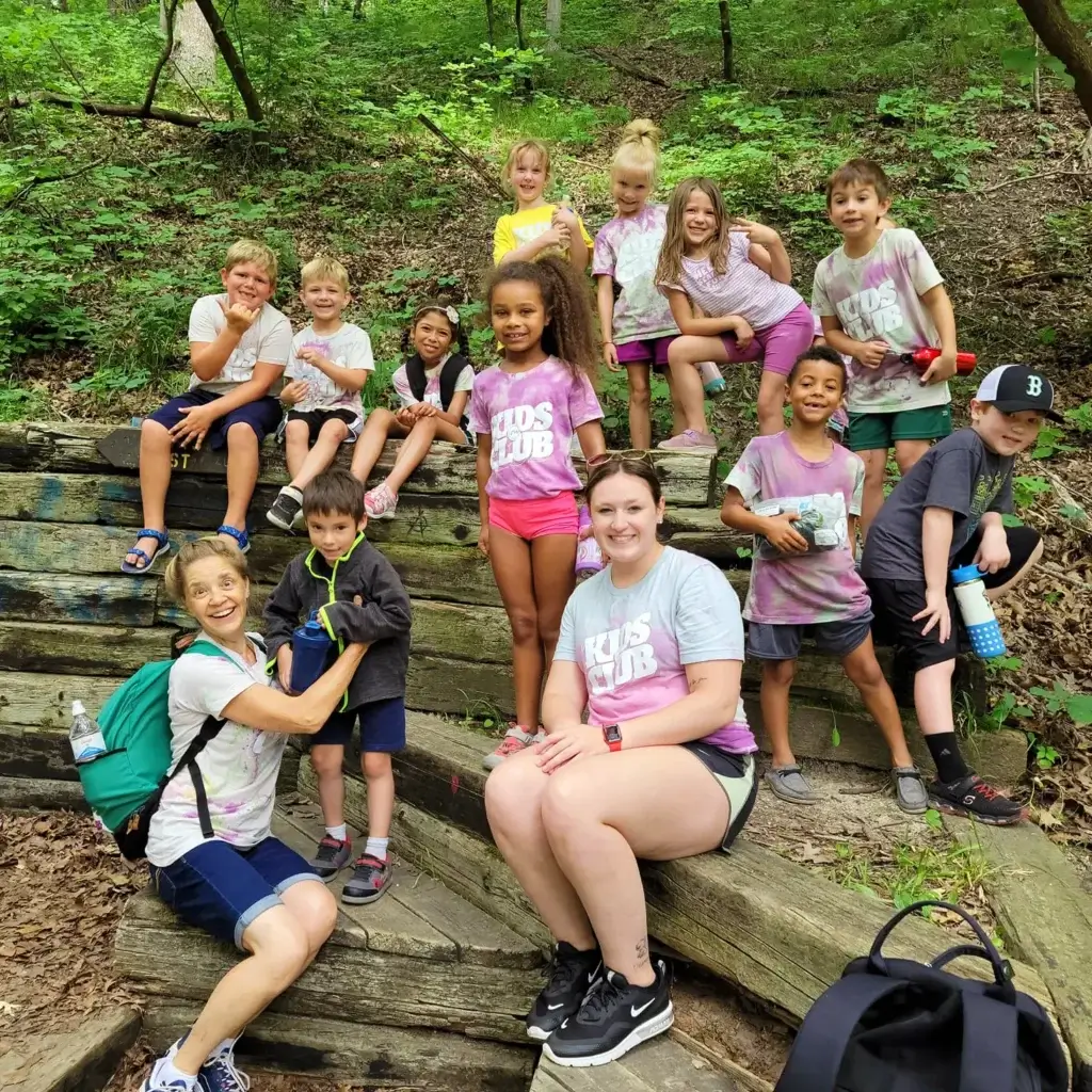 Two women and a group of kids smile and pose together on a forest trail.