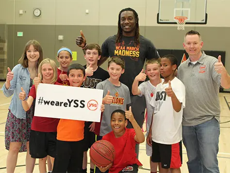 Adults and a group of children standing together in a gym and smiling while giving a thumbs up. One child holds a sign that says #WeAreYSS.