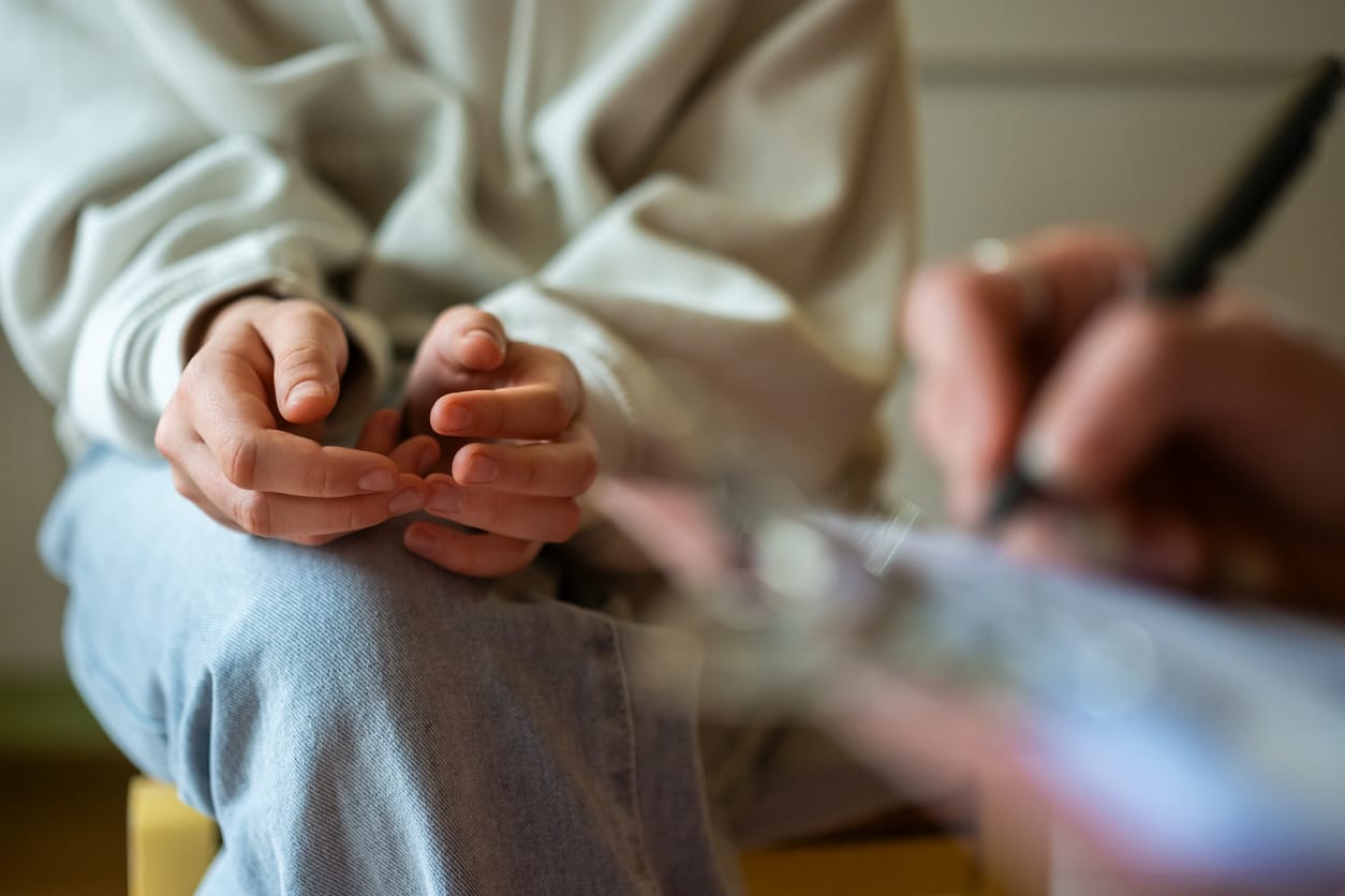 Close-up image of a teenage girl in casual clothes nervously fidgeting during a mental health therapy session with a professional taking notes.