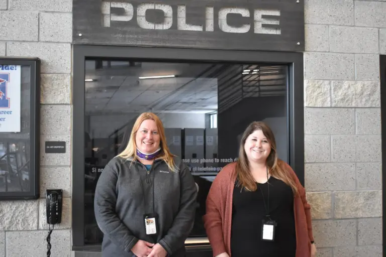 Two women stand and smile in front of a window with a police sign above their heads.